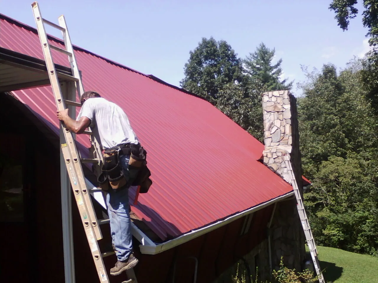 Roofer on a ladder beside a red metal roof and stone chimney — Soffit & Fascia Repair by Roofing Brothers in Harwood Heights